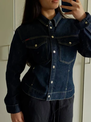 Mörkblå jeansjacka med fickor - Snygg mörkblå jeansjacka med markerade sömmar och två bröstfickor med knappar. Klassisk krage och silverfärgade knappar framtill. Figurnära passform som ger en cool siluett. Perfekt att styla med både byxor och kjol.