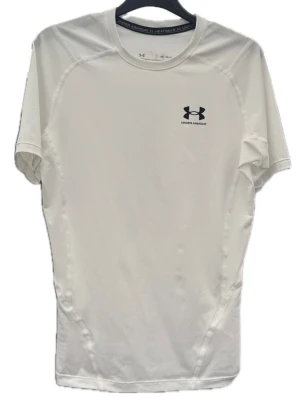 Vit tränings-tshirt från Under Armour - Snygg vit tränings-tshirt från Under Armour med korta ärmar och svart logga på bröstet. Tillverkad i ett lätt och funktionellt material som andas, perfekt för gymmet eller löpning. Sitter tight och har sömmar som ger en sportig look. Liten tråddragning på armen