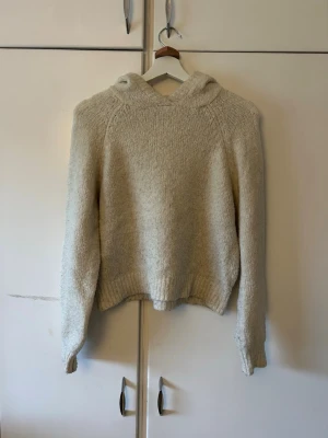 Beige stickad hoodie - Mysig beige stickad hoodie med luva och långa ärmar. Tröjan har en avslappnad passform och ribbstickade muddar vid ärmslut och nederkant. Perfekt för kalla dagar när du vill vara både varm och stilren.