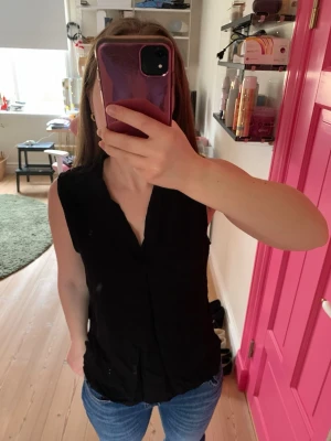 Svart ärmlös blus med v-ringning - Snygg svart ärmlös blus med v-ringning och enkel, stilren design. Perfekt att matcha med jeans eller kjol för en clean look. Tillverkad i mjukt tyg som känns skönt mot huden. Står storlek L på lappen, men passar storlek M och S. 
