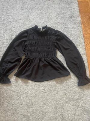 Svart smockad blus med volangkrage - Supersnygg svart blus med smockad överdel och volangkrage. Långa puffiga ärmar med resår vid ärmslut och lätt utsvängd nederdel. Perfekt till jeans eller kjol för en trendig look.