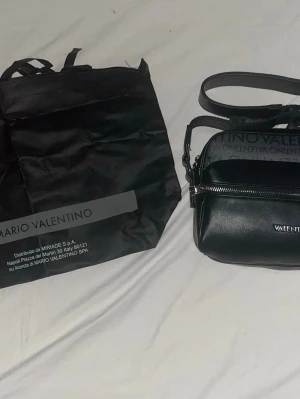 Svart axelväska från Mario Valentino - Snygg svart axelväska från Mario Valentino med justerbar axelrem och silverfärgad dragkedja. Väskan har logotyp framtill och ett extra fack med tryckt Valentino-mönster upptill. Tillverkad i konstläder och levereras med original dustbag.