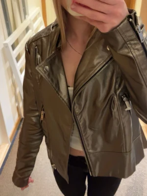 Metallic brun skinnjacka med dragkedjor - Snygg metallic brun skinnjacka i bikerstil med silvriga dragkedjor och detaljer. Kort modell med snedställd dragkedja framtill och fickor med dragkedja. Perfekt för att lyfta vilken outfit som helst.
