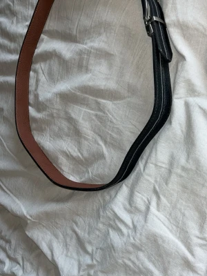 Svart och brunt läderbälte - Snyggt bälte i svart läder med brun insida och silverfärgat metallspänne. Bältet har en klassisk design med stilrena sömmar längs kanterna. Perfekt accessoar för att lyfta vilken outfit som helst.