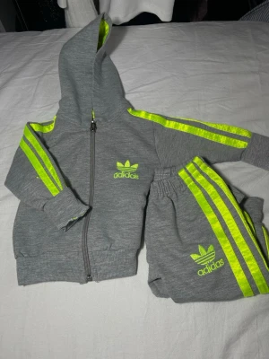 Grått  mjukisset med neongula detaljer - Säljer ett grått loungewear-set med hoodie och byxor. Setet har neongula ränder längs ärmar och ben samt Adidas logga broderad på både tröja och byxa. Mjukt material och ribbade muddar för extra komfort. Perfekt för chill dagar hemma. Storlek 68 bebis.