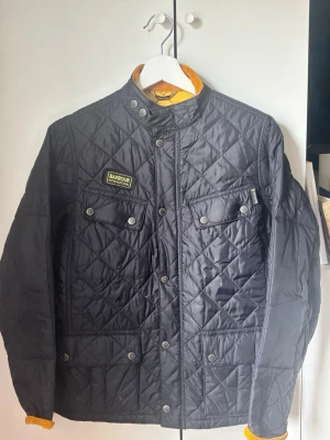  Barbour jacka - Säljer en svart quiltad jacka från Barbour med tryckknappar, två bröstfickor och två sidofickor. Jackan har ståkrage med tryckknapp och gul kontrastfärg på insidan av kragen och ärmsluten. Klassisk Barbour-logga på bröstet. Perfekt för dig som gillar stilren och tidlös design.