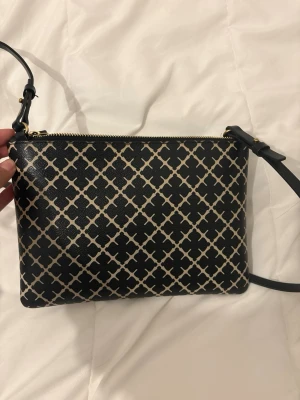  Malene Birger axelväska - Snygg svart axelväska från By Malene Birger med klassiskt beige mönster och guldfärgad dragkedja. Väskan har justerbar axelrem och är tillverkad i tåligt konstläder. Perfekt för dig som gillar stilrena accessoarer med en twist. Utan några defekter 