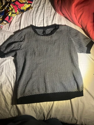 Svartvit mönstrad topp  - Säljer en svartvit topp med zigzag-mönster. Toppen har korta ärmar, rund halsringning och ribbade muddar vid ärmslut och nederkant. 