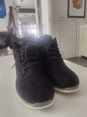Svarta kängor från Everest - Säljer ett par svarta kängor från Everest med vita sulor och gråa skosnören. Kängorna har en sportig design med högre skaft och är tillverkade i syntetmaterial. Perfekta för dig som vill ha ett stilrent och bekvämt alternativ till vardags.