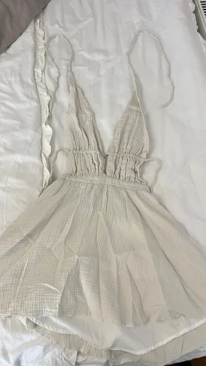 Vit omlottklänning med knytband - Söt vit kortklänning med omlottdesign och smala knytband. Klänningen har djup v-ringning, rynkad midja och är tillverkad i ett lätt, luftigt bomullstyg. Perfekt för varma dagar och ger en avslappnad men trendig look.