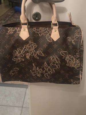 Väska  - Säljer en brun handväska  med klassiskt monogrammönster och ljusa handtag. Väskan har blommiga detaljer i beige och guld, samt guldfärgade metalldetaljer. Tillverkad i slitstarkt läder med två korta handtag och dragkedja upptill.