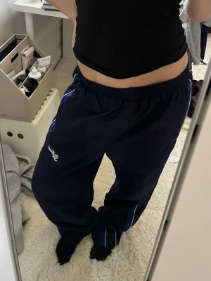 Mörkblå trackpants med blå detaljer - Mörkblå byxor från Siforever med blå och vita ränder längs benen. Byxorna har resår i midjan och en avslappnad passform. Tillverkade i ett lätt och bekvämt material, perfekta för en sportig stil. Logga på vänster lår.