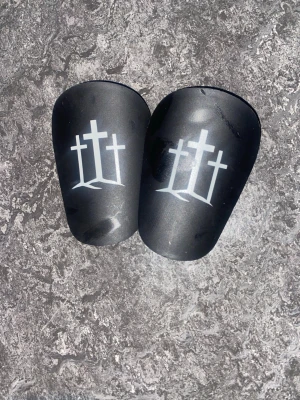 Svarta benskydd med korsmotiv - Säljer ett par svarta benskydd med vitt tryck av tre kors på framsidan. Benskydden har en klassisk form och är tillverkade i ett hårt plastmaterial som ger bra skydd vid fotboll eller andra sporter.