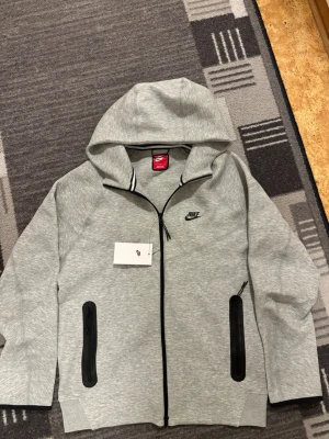 Grå Nike hoodie med dragkedja - Snygg grå hoodie från Nike med svart logga på bröstet och hel dragkedja framtill. Två fickor med svarta dragkedjor och en stor huva. Tillverkad i mjukt sweatshirtmaterial som känns skönt mot huden. Perfekt för en avslappnad streetwear-look.