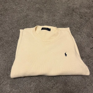 Beige stickad tröja från Polo Ralph Lauren - Klassisk beige stickad tröja från Polo Ralph Lauren med rund halsringning och diskret blå logga broderad på bröstet. Tröjan har långa ärmar och ribbade muddar vid ärmslut och nederkant. Perfekt för en stilren och avslappnad look. OBS, storleken är M men skulle säga den passar S-M