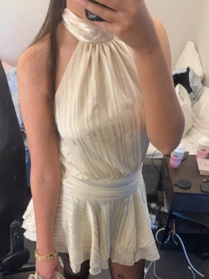 Krämvit halterneckklänning - Snygg krämvit klänning med halterneck och subtila ränder i tyget. Klänningen har markerad midja och ett kort, utsvängande kjolparti som ger en feminin siluett. Perfekt för dig som gillar en chic och trendig look.