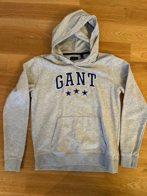 Hoodie med tryck - Snygg grå hoodie från GANT med blått broderat logotryck och tre stjärnor på bröstet. Tröjan har en stor huva, ribbade muddar och en klassisk magficka. Materialet är mjukt och bekvämt, perfekt för chill dagar. Passar dig som gillar stilren design och kvalitetskläder.