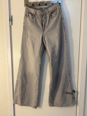 Gråa vida jeans med hög midja - Säljer ett par ljusgrå jeans med hög midja och riktigt vida ben. Klassisk femficksmodell med dragkedja och knapp framtill. Jeansen har en avslappnad passform och är tillverkade i denim. Perfekta för en trendig och chill look.