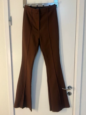 Bruna utsvängda kostymbyxor - Säljer ett par bruna kostymbyxor med hög midja och utsvängda ben. Byxorna har pressveck framtill och slitsar nedtill på benen för en trendig look. Perfekta för dig som gillar retrostil och vill sticka ut lite extra.