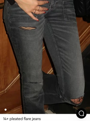 Grå slitna bootcut jeans - Säljer ett par grå bootcut jeans med slitningar vid knäna och låren. Jeansen har låg midja, vida ben och med några hål på benen, säljer för att de inte kommer till användning och dem är nästan helt nya! Skriv för mer bilder.