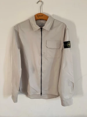 Stone Island Overshirt - Stilren beige overshirt från Stone Island med svart dragkedja framtill, klassisk krage och en bröstficka med lock. Ikonisk svart patch med logga på vänster ärm. Tillverkad i ett lätt, slätt material som ger en clean look. Perfekt för lager-på-lager och streetwear.