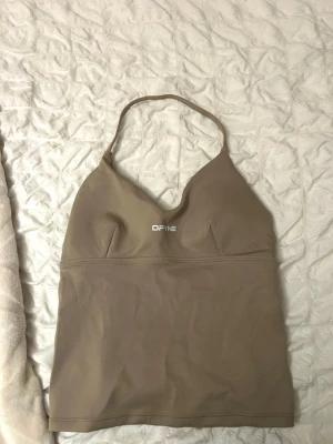 DFYNE Hannah Pearson halter neck top  - Säljer en beige linnetopp från DFYNE med smala axelband och v-ringad hals. Toppen har en stilren design med logga framtill och är tillverkad i ett stretchigt syntetmaterial som passar perfekt till träning. Aldrig använd 