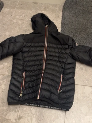 Svart dunjacka med huva från Moncler - Snygg svart dunjacka från Moncler med huva och två dragkedjeförsedda fickor. Jackan har Moncler-logga på ärmen och coola detaljer med röd, vit och blå dragkedja. Perfekt för kalla vinterdagar och riktigt stilren.