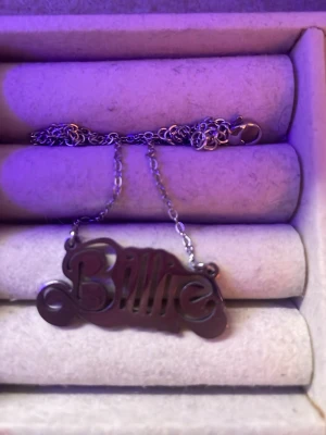 Billie eilish halsband - Unikt halsband i silverfärg med ett hängsmycke där det står 'Billie' i en cool, snirklig font. Kedjan är tunn och smidig med klassiskt lås. Perfekt accessoar för dig som vill sticka ut med ett personligt smycke.