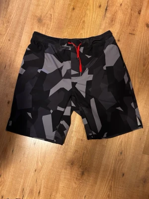 Svarta träningsshorts  med camomönster - Säljer ett par svarta träningsshort från ICANIWILL med grått camomönster och röd dragsko i midjan. Materialet är snabbtorkande och perfekt för träning utomhus eller inomhus. Snygg detalj med röd logga på benet. Sköna och stilrena träningsshort för dig som gillar sportig look.