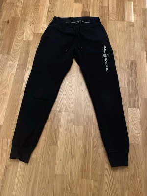 Svarta Sail Racing mjukisbyxor M - Svarta mjukisbyxor från Sail Racing med logga längs benet. Byxorna har resår i midjan med dragsko, ribbade muddar vid benslut och mjukt fleecefoder på insidan. Perfekta för chill eller sport. Snygg och enkel design.
