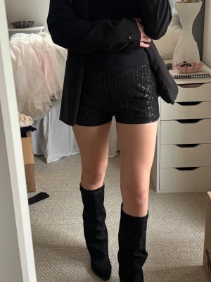 Svarta glittriga shorts med paljetter - Snygga svarta shorts med glittriga paljetter och hög midja. De har en tight passform och är perfekta för att sticka ut. Matcha med boots eller klackar för en trendig look.