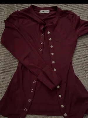 Vinröd långärmad kofta från Sister S - Stilren vinröd kofta från Sister S med långa ärmar och dekorativa silverfärgade knappar längs hela framsidan. Koftan har en mjuk och tunn kvalitet, perfekt för lager på lager och en snygg detalj till din outfit.