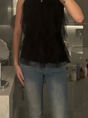 Svart volang topp med meshdetaljer - Säljer en svart ärmlös topp med volanger och meshdetaljer. Toppen har en hög halsringning dekorerad med små svarta paljetter.