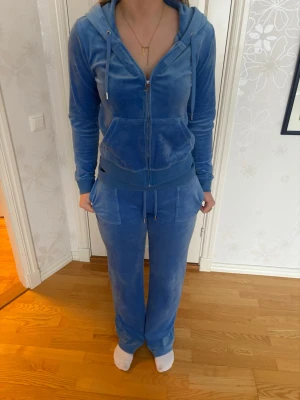 Blå velour mjukisbyxor Juicy Couture - Säljer ett par blå velour mjukisbyxor från Juicy Couture. Byxorna har dragsko i midjan, raka ben och stora fickor framtill. Perfekta för en chill och avslappnad stil. Matcha med hoodie för en komplett look.