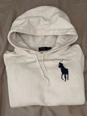Vit hoodie från Polo Ralph Lauren - Snygg vit hoodie från Polo Ralph Lauren med stor marinblå logga broderad på bröstet. Klassisk modell med huva och dragsko samt ribbade muddar vid ärmslut och midja. Perfekt för en avslappnad och stilren look. Jätte liten fläck på luvan samt en jätte litet hål i armen