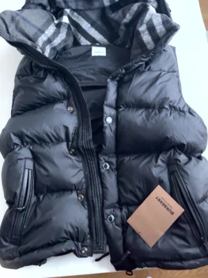 Burberry vest/jacka  - Kan gå ner o pris isåfall buda. Snygg svart dunväst/jacka från Burberry med klassiskt rutigt foder i huvan. Västen har dragkedja och knappar framtill, två fickor med dragkedja och en avtagbar huva. Perfekt för lager på lager och riktigt varm tack vare dunfyllningen. 