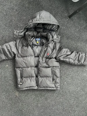 Svart dunjacka från Polo Ralph Lauren - Snygg svart dunjacka från Polo Ralph Lauren med huva och dragkedja framtill. Jackan har två fickor med dragkedja och den klassiska röda loggan broderad på bröstet. Perfekt för kalla vinterdagar och riktigt stilren look.