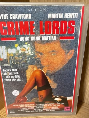 Crime Lords - Hong Kong Maffian VHS - Actionfilm på VHS med titeln 'Crime Lords - Hong Kong Maffian' med Wayne Crawford och Martin Hewitt i huvudrollerna. Omslaget har röd och blå färg med stora bokstäver och motiv från filmen. Perfekt för samlare av äldre actionfilmer.