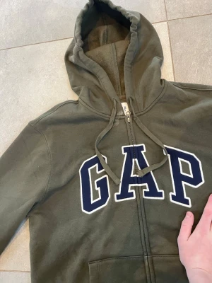 Olivgrön zip hoodie från GAP - Snygg olivgrön hoodie från GAP med dragkedja och stor logga i vitt och marinblått på bröstet. Hoodien har huva med dragsko och känguruficka framtill. Tillverkad i mjukt sweatshirtmaterial som är perfekt för chill dagar.