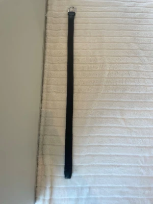 Svart flätat bälte från JOHN HENRIC - Snyggt svart flätat bälte med ett stilrent silverfärgat metallspänne. Bältet har en enkel och clean look som passar till de flesta outfits. Materialet är mjukt och flexibelt, vilket gör det bekvämt att bära hela dagen.