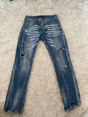 Dsquared2 blå slitna jeans - Säljer ett par blå jeans från Dsquared2 med slitningar, hål och färgstänk i vitt och rött. Jeansen har rak passform, klassisk femficksdesign och läderpatch med röd logga bak. Perfekta för en edgy och trendig streetstyle-look.