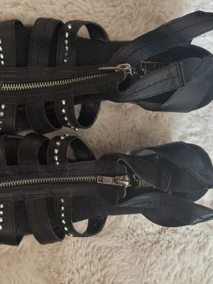 Svarta sandaler med nitar och dragkedja - Säljer ett par svarta sandaler med chunky klack, dragkedja framtill och coola nitar längs remmarna. Skorna har flera breda remmar i konstläder och en stabil träfärgad klack. Perfekta för dig som gillar edgy detaljer och vill sticka ut.