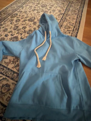 Synda world hoodie - Säljer en ljusblå hoodie med stor huva och vita, tjocka dragsnören. På ena ärmen finns en broderad text i vitt som ger en unik touch. Hoodien har en klassisk magficka och är gjord i mjukt bomullsmaterial. Perfekt för chill dagar eller streetwear-stil.BILIIGT PRIS KOM PÅ ME BUD