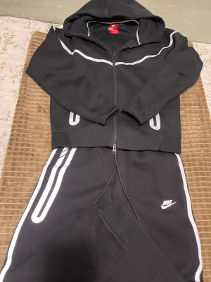 Svart Nike träningsset med huva - Snyggt svart träningsset från Nike med vit logga på byxan och vita detaljer längs ärmar och ben. Setet består av en zip-hoodie med dragsko och matchande byxor med resår och snörning i midjan. Perfekt för gymmet eller chill.