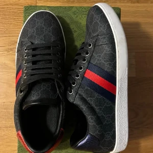 Gucci sneakers med röd/blå rand - Snygga svarta Gucci sneakers med monogrammönster och klassiska ränder i rött och blått på sidan. Skorna har vita sulor, svarta skosnören och detaljer i rött och blått på hälen. Tillverkade i canvas och läder för en exklusiv känsla.