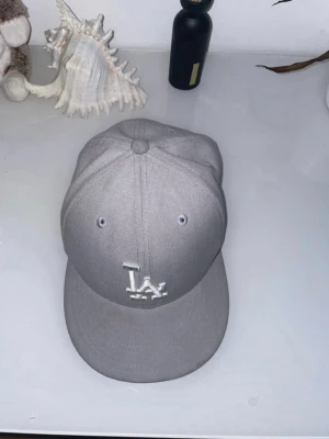 Grå LA-keps med broderad logga - Snygg grå keps från New Era med LA-logga broderad framtill i vitt. Klassisk modell med böjd skärm och ventilationshål. Perfekt accessoar för en sportig och trendig look.