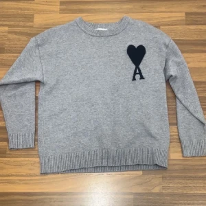 Grå stickad tröja från Ami Paris - Snygg grå stickad tröja från Ami Paris med svart hjärta och bokstaven A broderat på bröstet. Tröjan har rund halsringning, ribbade muddar och nederkant. Perfekt för dig som gillar stilrena och trendiga plagg.