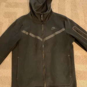 Svart Nike hoodie med dragkedja - Snygg svart hoodie från Nike med hel dragkedja och huva. Har diskreta gråa ränder över bröstet och ärmen samt Nike-logga på vänster sida. Praktisk ficka med dragkedja på ena ärmen. Perfekt för en sportig och avslappnad stil. Priset kan diskuteras 
