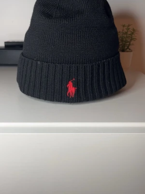 Svart stickad mössa från Polo Ralph Lauren - Svart ribbstickad mössa från Polo Ralph Lauren med klassisk röd broderad logga framtill. Mössan har uppvikt kant och är tillverkad i mjukt stickat material som håller dig varm under kalla dagar.