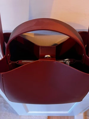 Vinröd handväska i läder🍷🍒 - Stilren vinröd handväska i slätt läder med breda handtag och en praktisk innerficka med dragkedja. Väskan har en enkel knappstängning och rymligt fack, perfekt för att bära dina essentials. Snygg och minimalistisk design som passar till många outfits.